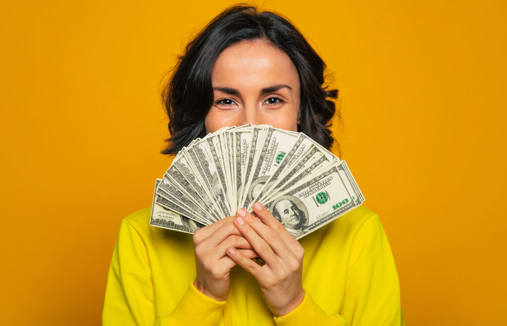 Close-up photo of a young girl in a yellow hoodie, smiling with  her eyes, hiding her face behind big amount of money in her hands.
