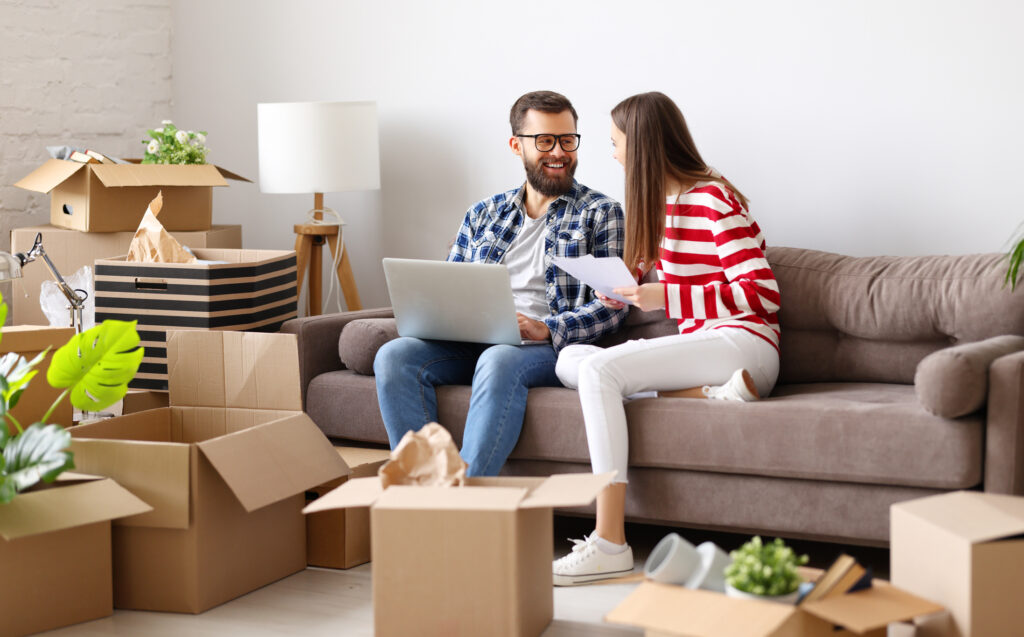 Happy young family couple with laptop and papers sitting on sofa in room with packed boxes and discussing insurance contract after relocation into new home