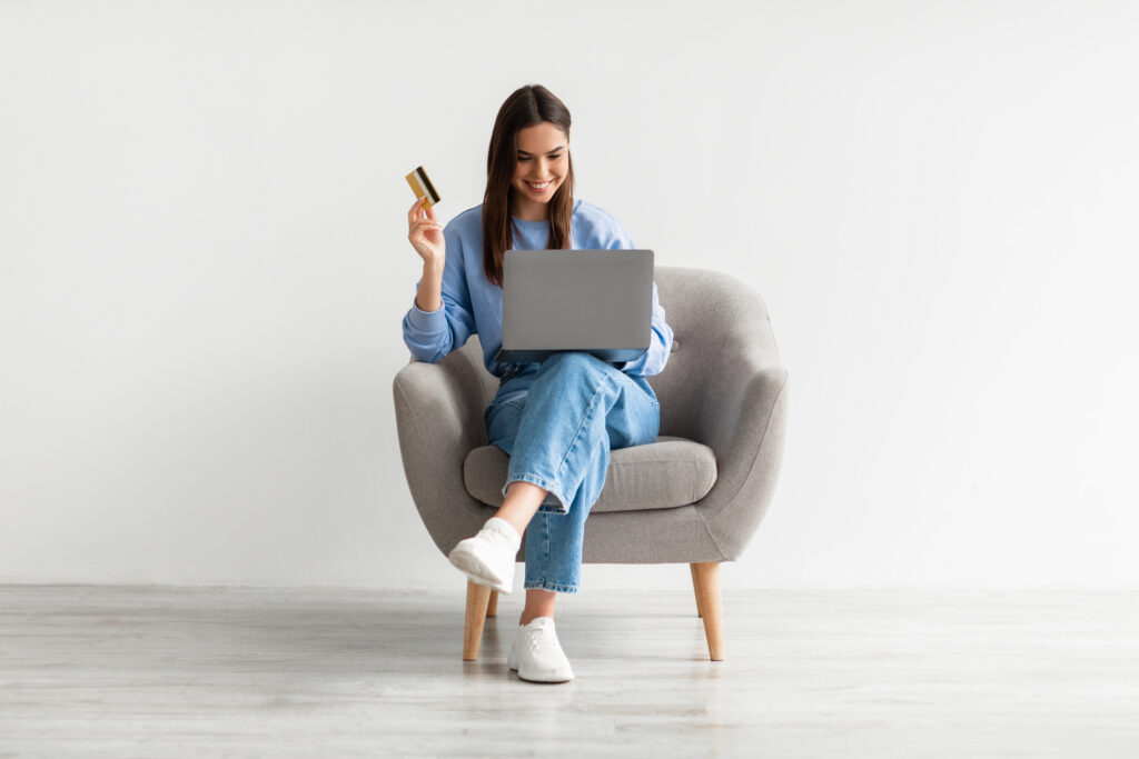 Lovely young woman sitting in armchair with laptop, using credit card to buy things on web
