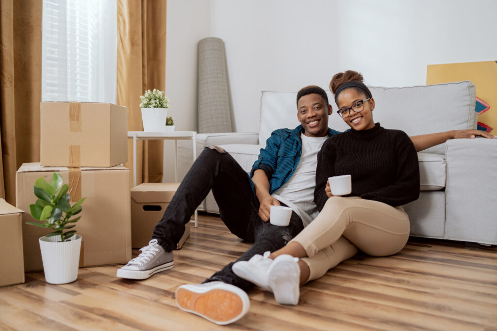 Smiling couple sitting on the floor relaxing after moving in, drinking coffee in their new apartment, around them boxes with unpacked things, the husband embraces his wife