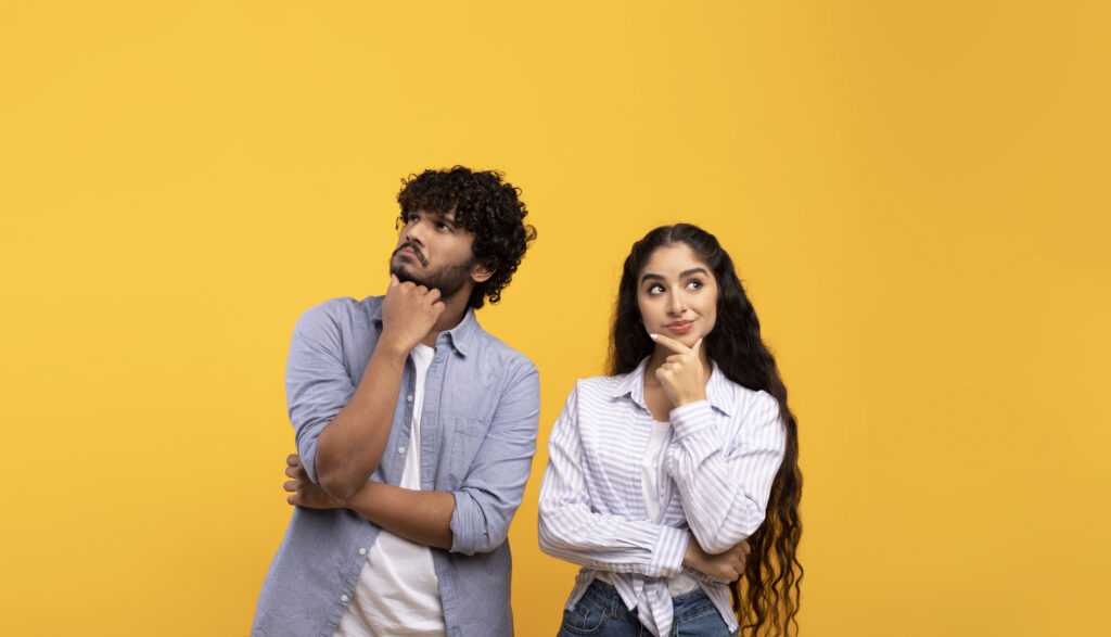 Portrait of pensive man and woman thinking and looking up at free space, touching chin, standing over yellow studio background. Thoughtful couple making decision