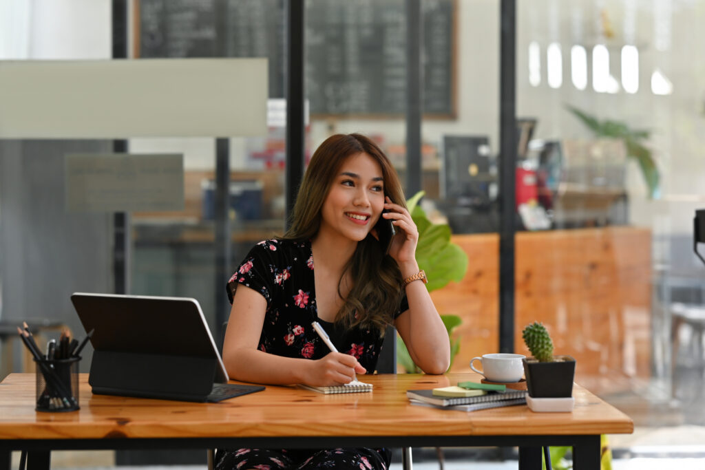 A portrait of a young woman sitting in the office using a telephone, laptop, working on the documents, insurance agent on the phone.