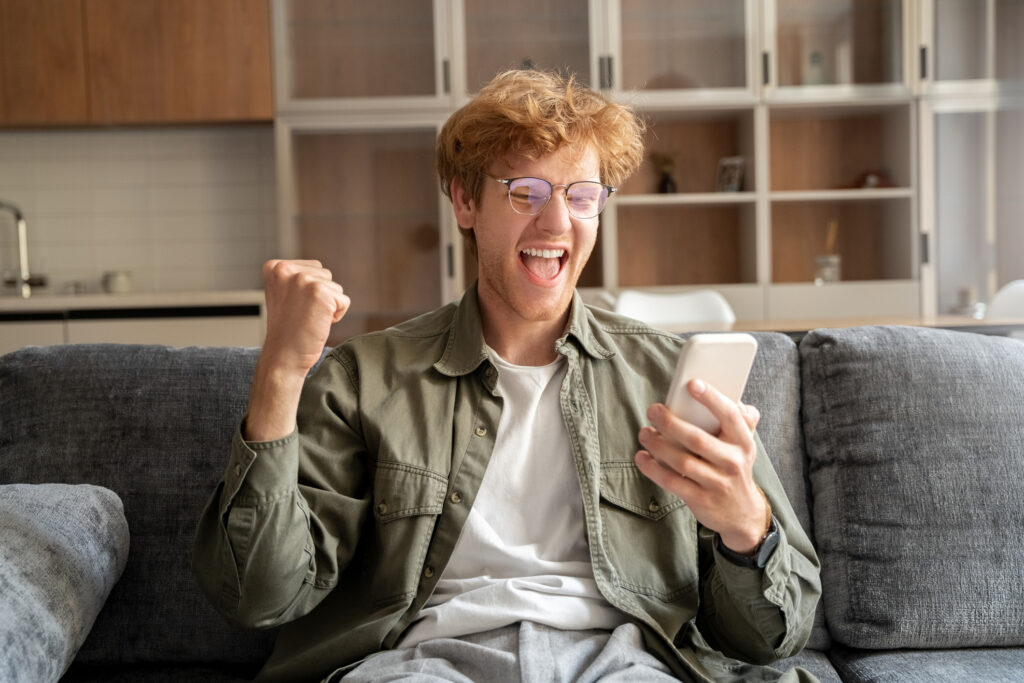 Happy overjoyed excited young  man in glasses gesturing and screaming yes to mobile phone. Unexpected reward or success, online investment victory