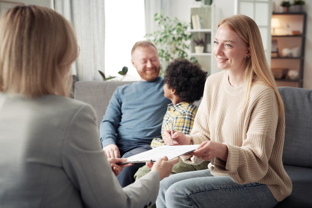 Happy young woman signing contract about adoption while talking to social worker with her family in background