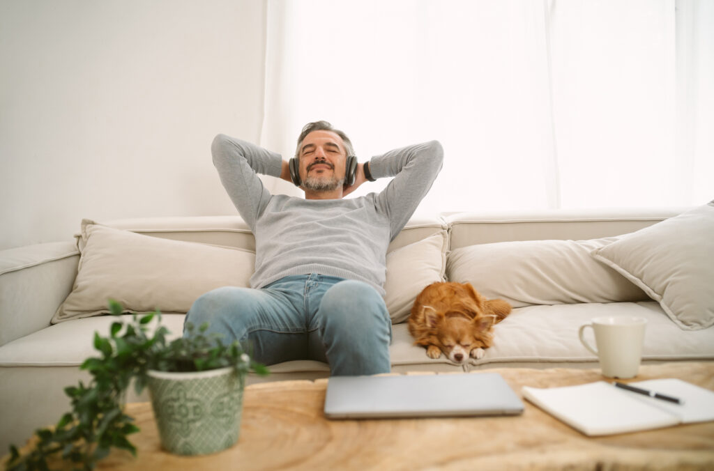 Calm man sitting on sofa listening to music enjoying meditation for sleep and peaceful mind in wireless headphones, leaning back with his lovely chihuahua dog sit besides.