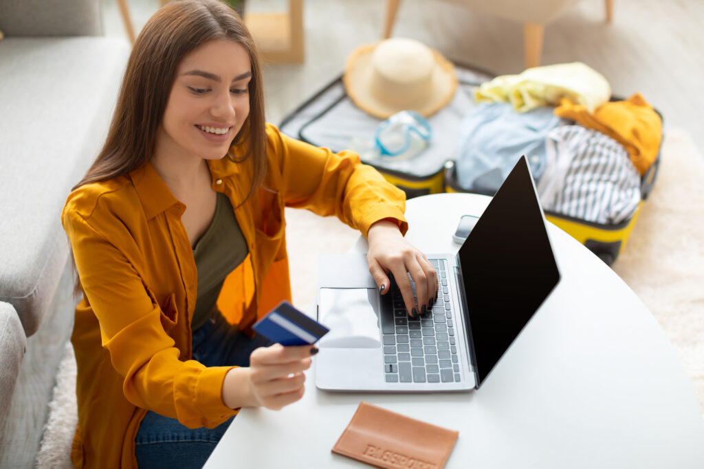 Cheery young lady using credit card and laptop booking trip, making hotel reservation on web from home