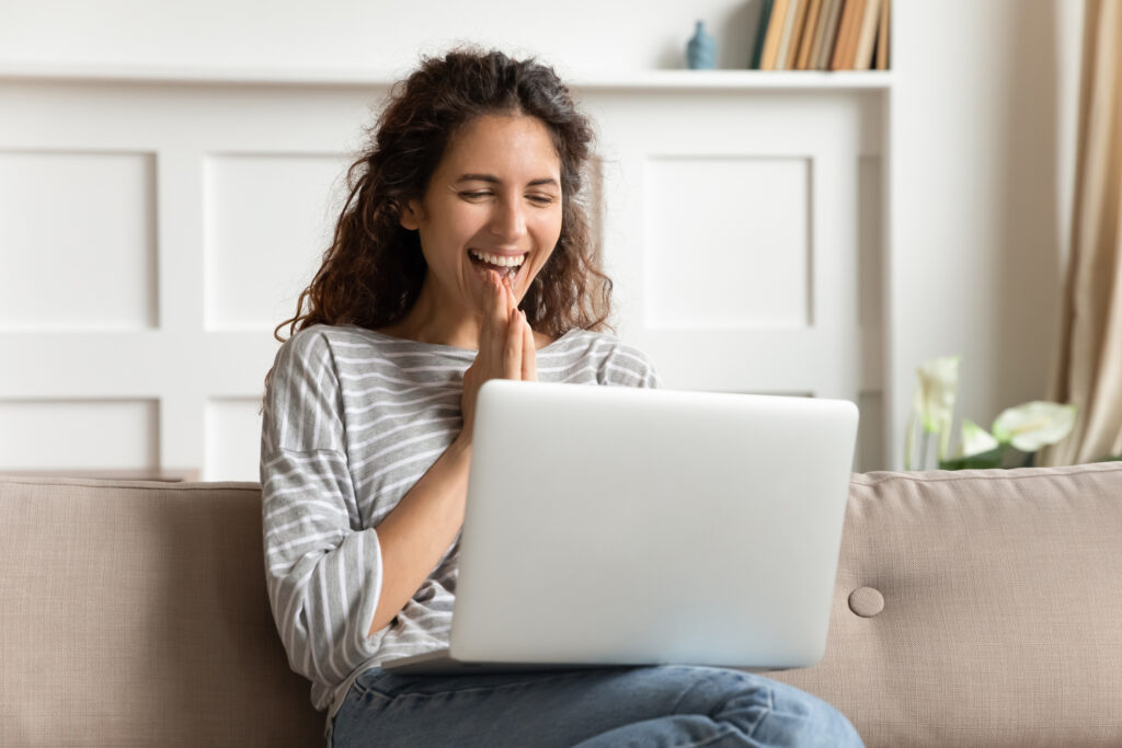 Overjoyed young woman looking at laptop screen, feeling excited about good news, sitting on sofa in living room. 