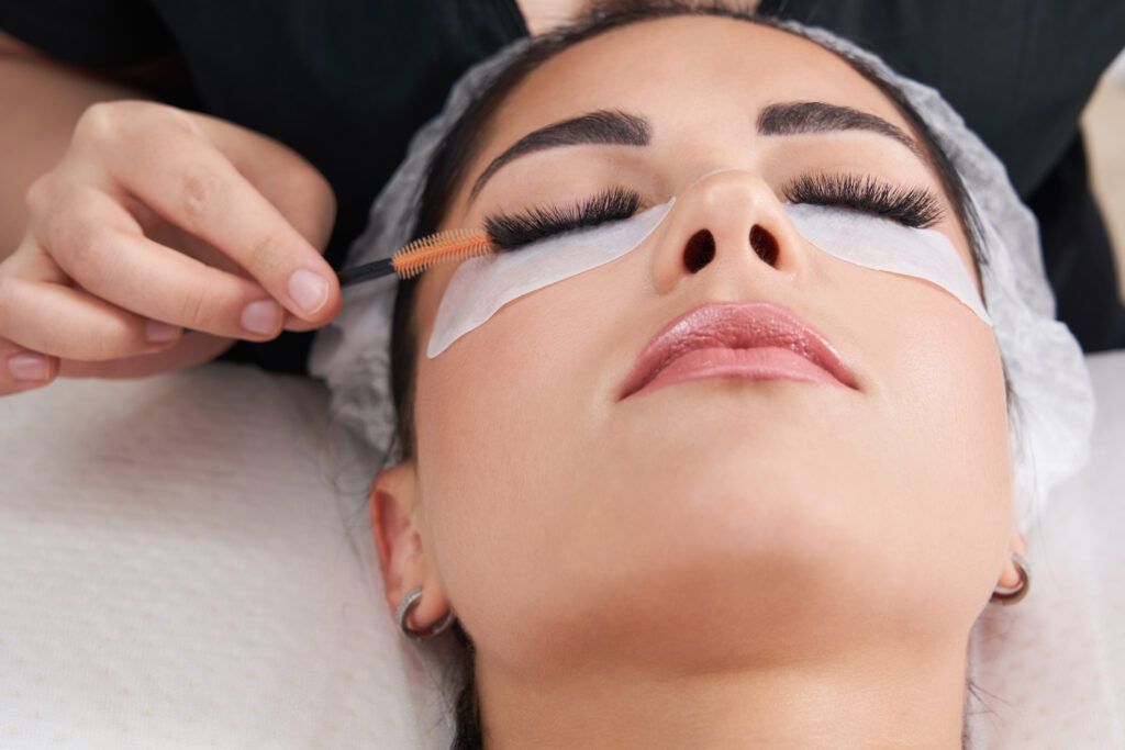 Cosmetologist making eyelash extension and correction using brush in salon