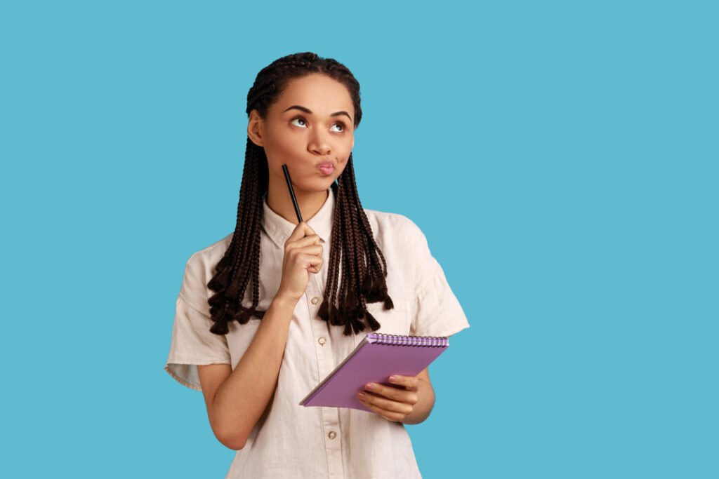 Portrait of pensive woman holding paper notebook, having thoughtful facial expression, planning, wearing white shirt. Indoor studio shot isolated on blue background. 