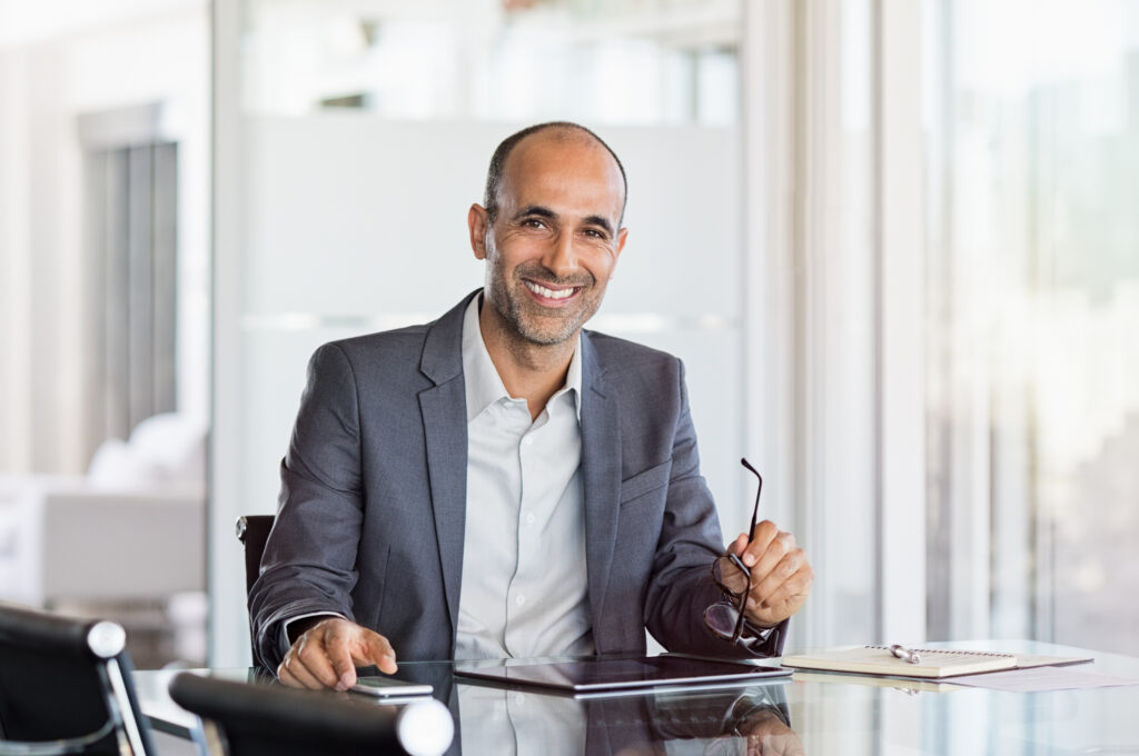 Happy mature financial professional man holding spectacles in modern office. Sitting in meeting room with phone and tablet. Smiling man in suit in a elegant office.