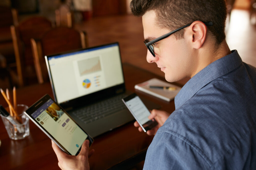 Attractive man in glasses working with multiple electronic devices. has tablet and smartphone in hands and laptop on table with charts on screen. Multitasking theme.