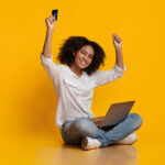 Online Cashback Concept. Portrait Of Happy Woman Celebrating Success With Laptop And Credit Card, Raising Hands In Excitement, Yellow Background