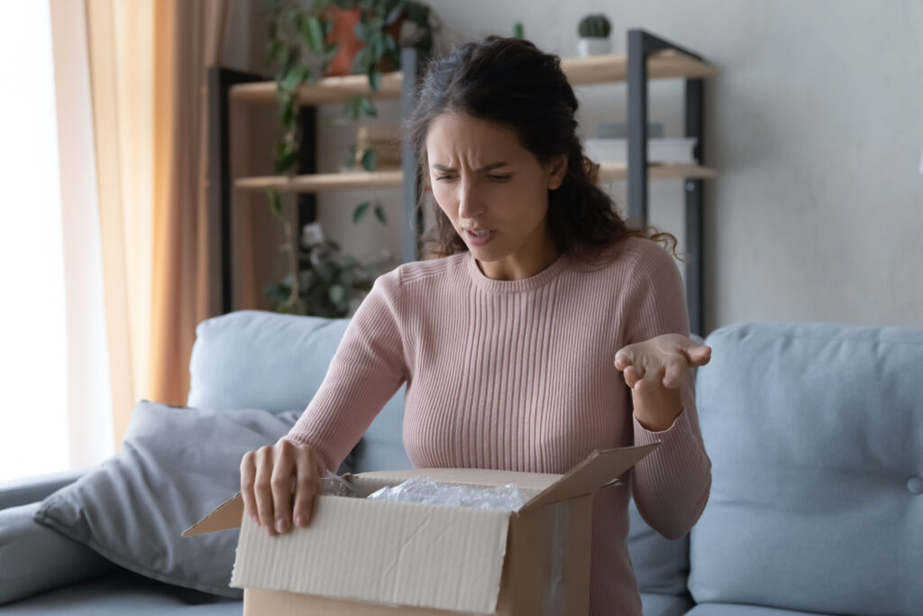 Woman unpacking parcel, wrong or broken online store order, sitting on couch at home, dissatisfied female looking in cardboard box, bad delivery service, displeased 