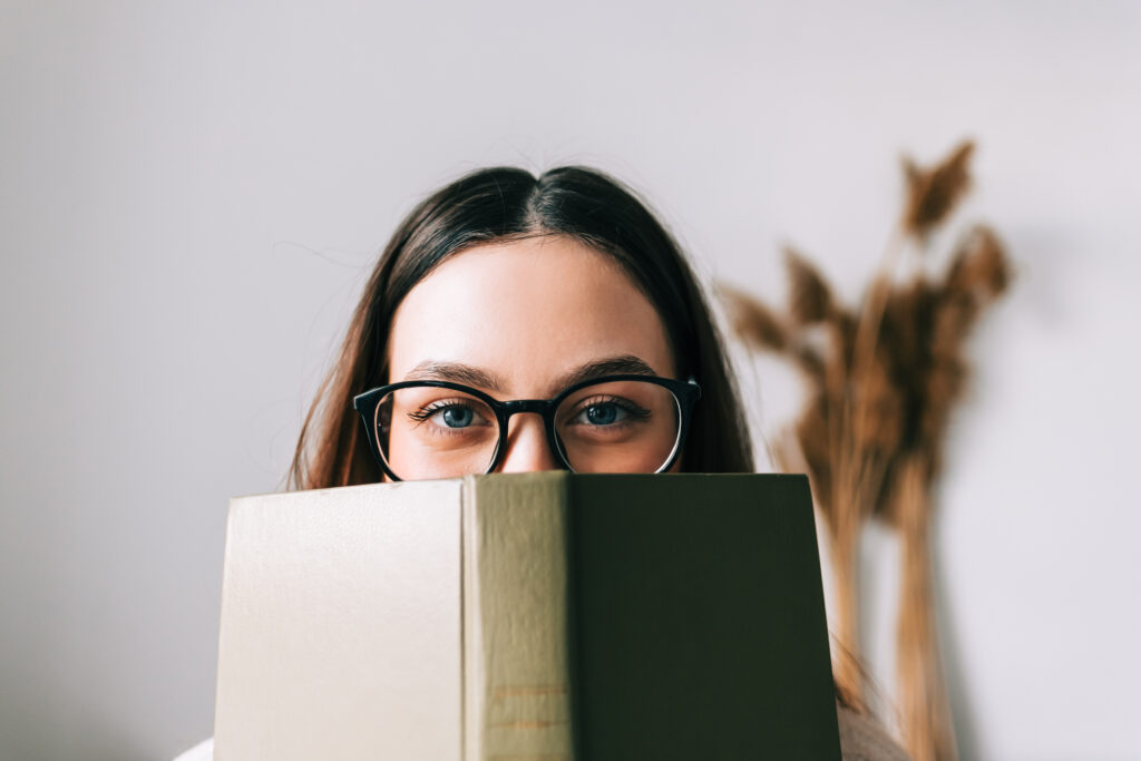 Portrait of young woman college student in eyeglasses hiding behind a book and looking at camera.