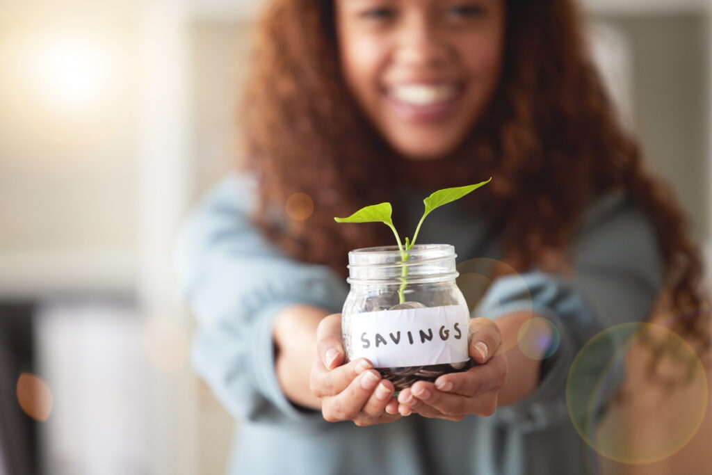 Young woman presenting her glass savings jar with a budding plant growing out from it at home. Happy  person smiling while planning, saving and investing for her future